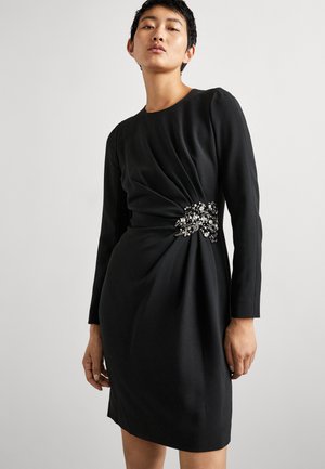 Abito lungo nero a maniche lunghe con silhouette aderente, dettaglio drappeggiato a torsione in vita e un adornamento decorativo di perline su un lato.