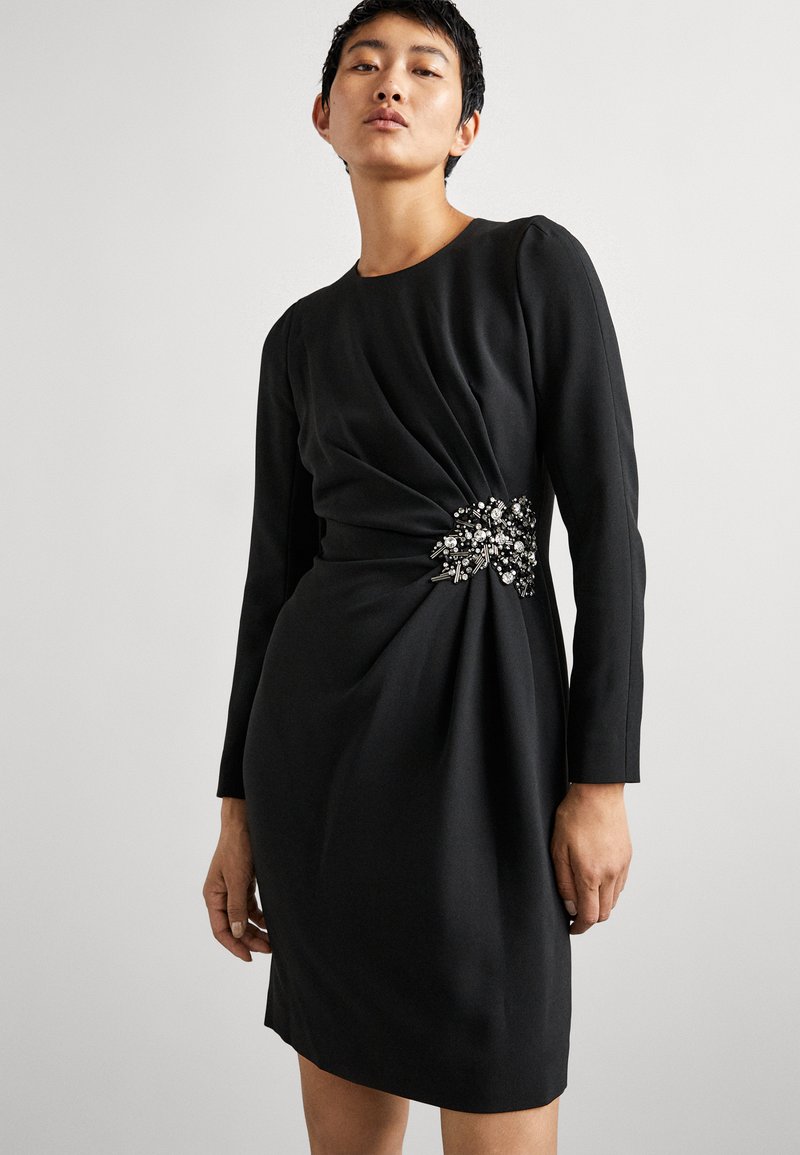 Abito lungo nero a maniche lunghe con silhouette aderente, dettaglio drappeggiato a torsione in vita e un adornamento decorativo di perline su un lato.