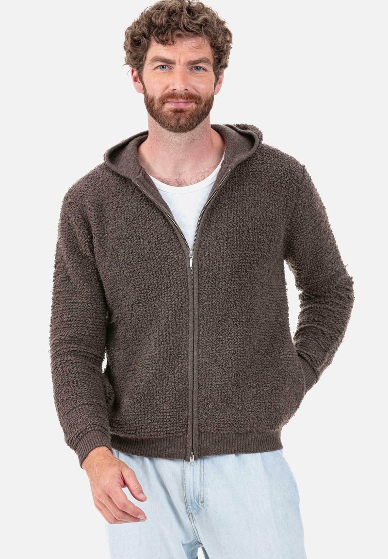 Veste à capuche zippée en polaire marron avec une surface texturée, des poignets et un ourlet côtelés, et une capuche à cordon. Portée par-dessus une chemise blanche.