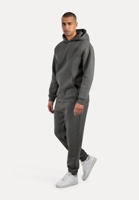 Sudadera gris con capucha y bolsillo tipo canguro, acompañada de pantalones de chándal gris a juego. Se lleva con zapatillas blancas, con detalles de logotipo sencillos.