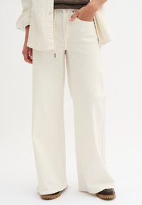 Pantaloni a gamba larga color avorio realizzati in tessuto morbido, con vita regolabile e tasche laterali. L'orlo è leggermente svasato, completando la vestibilità comoda.