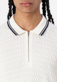 Biely textured polo s predným zipsom a navy pruhovaným golierom. Má pletený dizajn s jemným vzorom.