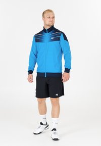 Blå idrottsjacka med svarta detaljer, dragkedja och ljusa ränder. Bärs med svarta shorts och vita sneakers, presenterad med varumärkesdetaljer.