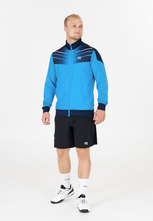 Blå idrottsjacka med svarta detaljer, dragkedja och ljusa ränder. Bärs med svarta shorts och vita sneakers, presenterad med varumärkesdetaljer.