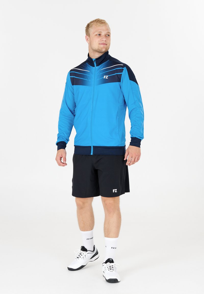 Blå idrottsjacka med svarta detaljer, dragkedja och ljusa ränder. Bärs med svarta shorts och vita sneakers, presenterad med varumärkesdetaljer.