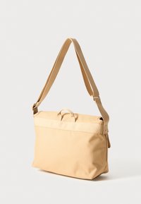 Beige lerretsveske med justerbar skulderrem, topphåndtak og enkel design. Har en jevn tekstur og rektangulær form med glidelås-lukking.