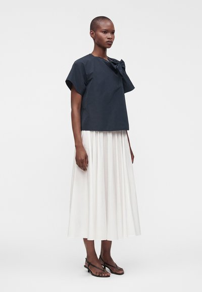 Femme portant un haut bleu marine à manches courtes avec un détail nœud et une jupe midi plissée blanche avec des talons noirs à brides, debout devant un fond uni.