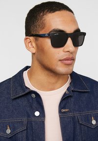 Ochelari de soare pătrați negri cu lentile închise la culoare, accent albastru pe brațe, purtați cu o jachetă din denim și o cămașă roz deschis.