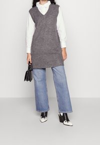 Nainen, jolla on yllään harmaa hihaton neuletunika valkoisen paidan päällä ja siniset farkut, pitää mustaa käsilaukkua ja käyttää mustavalkoisia korkokenkiä.