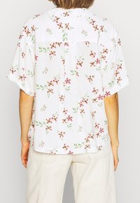 Personne portant un chemisier blanc à manches courtes avec des motifs floraux roses et un pantalon crème, vu de dos.