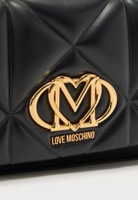 Bolso acolchado negro con un logotipo metálico dorado que presenta corazones entrelazados y la letra "M". Textura lisa con detalles brillantes.