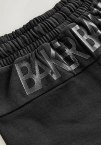 Zbliżenie na czarną tkaninę z elastycznym pasem i dużym błyszczącym napisem „BANGRBA” nadrukowanym u góry.