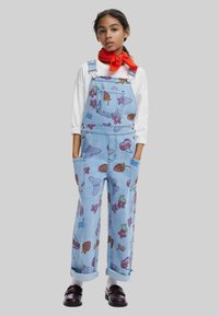 Salopette di jeans blu con motivi di frutta colorati, spalline regolabili e multiple tasche. Indossata con una camicia a maniche lunghe bianca.