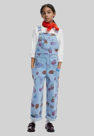 Salopette di jeans blu con motivi di frutta colorati, spalline regolabili e multiple tasche. Indossata con una camicia a maniche lunghe bianca.