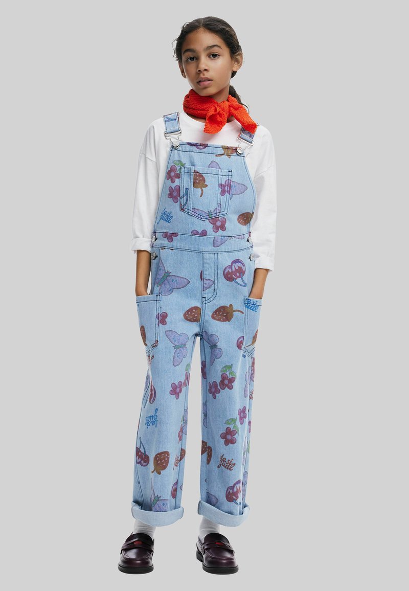 Salopette di jeans blu con motivi di frutta colorati, spalline regolabili e multiple tasche. Indossata con una camicia a maniche lunghe bianca.