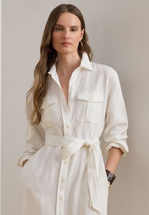 Γυναίκα με μακριά κυματιστά μαλλιά, φορώντας λευκό φόρεμα με κουμπιά και ζώνη στη μέση, με γυρισμένα μανίκια, μπροστινές τσέπες και σκούρο βραχιόλι καρπού.