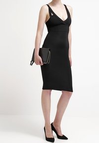 Schwarzes ärmelloses Kleid mit tiefem V-Ausschnitt und ausgeschnittenem Schulterdesign. Figurbetonte Silhouette, die bis zur Mitte der Wade reicht. Kombiniert mit einer kleinen schwarzen Clutch und hohen Schuhen.