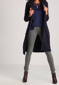 Cappotto lungo navy sopra una blusa navy con dettagli in pizzo, jeans skinny verde oliva e stivaletti bordo. Tessuto morbido e design aderente.