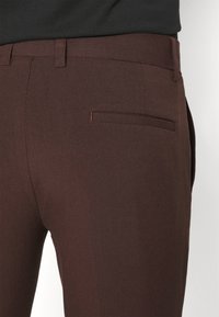 Bruna skräddarsydda byxor med en jämn yta, slim fit och en enda bakficka med röd söm som accent.