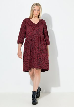 Robe bordeaux à imprimé léopard noir, décolleté en V, manches 3/4 et taille froncée. Portée avec des bottes noires à lacets. Texture lisse.