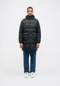 Giacca a vento nera con colletto alto, chiusura con zip e due tasche anteriori. Abbinata a jeans blu larghi e scarpe da ginnastica bianche.