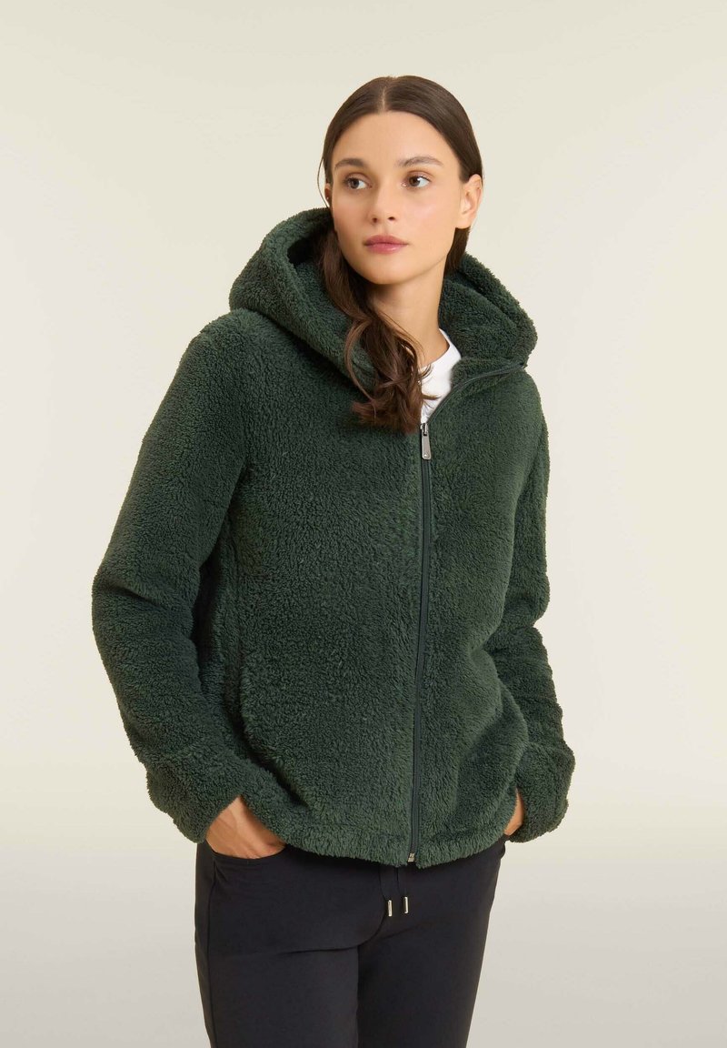 Giacca verde fuzzy con cappuccio e chiusura con zip, caratterizzata da una consistenza morbida e due tasche laterali, indossata su una maglietta bianca e abbinata a pantaloni scuri.