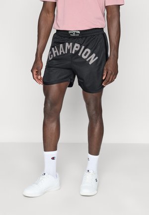 Fekete sportos rövidnadrág „CHAMPION” szürke felirattal, gumis derékrésszel, oldalzsebekkel, fehér zoknival és sportcipővel viselve.