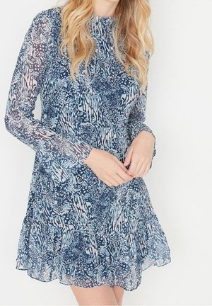 Mujer con cabello rubio largo que lleva un vestido azul con estampado, de manga larga y dobladillo con volantes, frente a un fondo blanco liso.