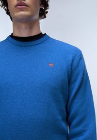 Blå stickad tröja med en texturerad yta, med rund halskrage och liten röd och vit flagg-emblem på bröstet.