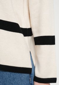Kremowy sweter z czarnymi paskami, o miękkiej fakturze, luźnym kroju i bocznym rozcięciu. Połączony z dżinsami.
