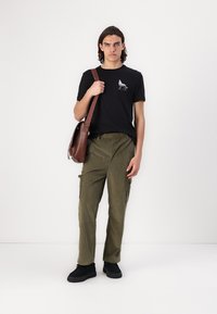 Zwarte katoenen t-shirt met wolf afbeelding, olijfgroene corduroy cargobroek en zwarte instap schoenen; bruine schoudertas met band.