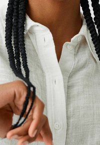 Biała lniana koszula z zapięciem na guziki, wykonana z teksturowanej tkaniny. Widać czarne splecione włosy, a ręka chwyta jeden z warkoczy.