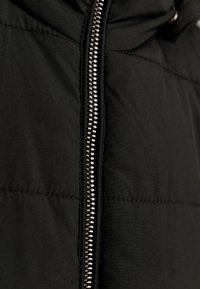 Zbliżenie na czarną pikowaną kurtkę z centralnym srebrnym metalowym zamkiem błyskawicznym i widocznym poziomym przeszyciem na tkaninie.