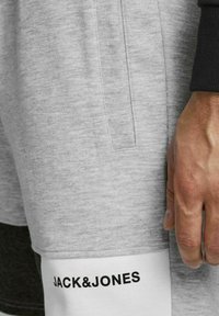 Šedé bavlněné šortky mají hladkou texturu, dvě boční kapsy a kontrastní černý pas. Značka "JACK&JONES" je vytištěna tučným černým písmem na lemu.