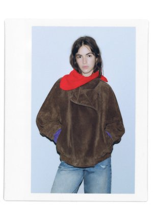 Une personne porte une veste marron oversize, une écharpe rouge vif et un jean bleu sur un fond de studio bleu pâle.