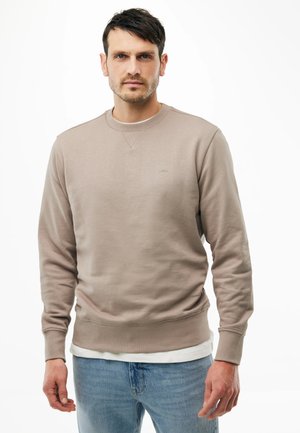 Hombre con cabello corto y oscuro, vestido con sudadera beige con cuello redondo y jeans azul claro, de pie frente a un fondo blanco.