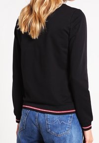 Una donna con lunghi capelli biondi indossa un top nero a maniche lunghe con bordi a strisce rosse e bianche, abbinato a jeans in denim blu, vista da dietro.