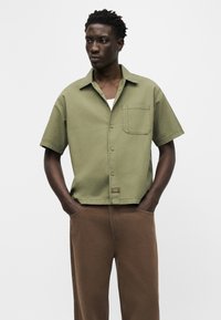 Νέος άνδρας που φοράει πουκάμισο με κοντά μανίκια σε ελιά πράσινο χρώμα και καφέ παντελόνι, στέκεται με τα χέρια στις τσέπες σε απλό φόντο.