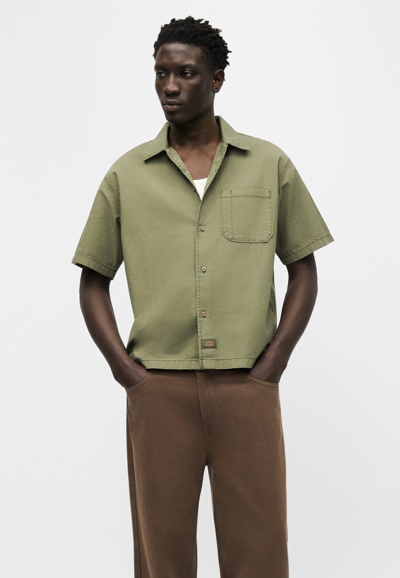 Joven con camisa de manga corta verde oliva abotonada y pantalones marrones, de pie con las manos en los bolsillos frente a un fondo sencillo.