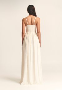 Maxi-jurk in off-white stof met een smock-borstvoeding, dunne bandjes en een vloeiende rok. De textuur is zacht en heeft een luchtige uitstraling.