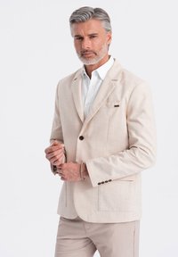 Blazer de lino beige con solapas de muesca, cierre de un solo botón, dos bolsillos de parche y una textura sutil. Combinado con una camisa blanca.