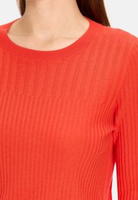 Haut tricoté rouge à côtes, doté d'un col rond et d'une coupe ajustée. La texture est lisse avec des rayures verticales et une épaisseur moyenne.