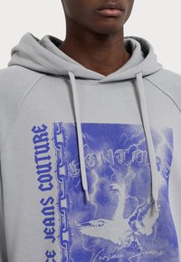 Felpa grigia chiaro con cappuccio, dotata di un design grafico blu di catene e un uccello, tessuto testurizzato e cordini regolabili.
