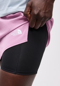 Κοντινό πλάνο ενός σκουρόχρωμου χεριού που σηκώνει μία ροζ φούστα The North Face, αποκαλύπτοντας μαύρα εφαρμοστά σορτς από κάτω.
