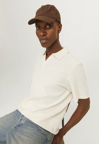 Polo blanco de canalé con cuello en V y mangas cortas, combinado con vaqueros de denim azul claro. Gorro marrón en la cabeza del modelo.