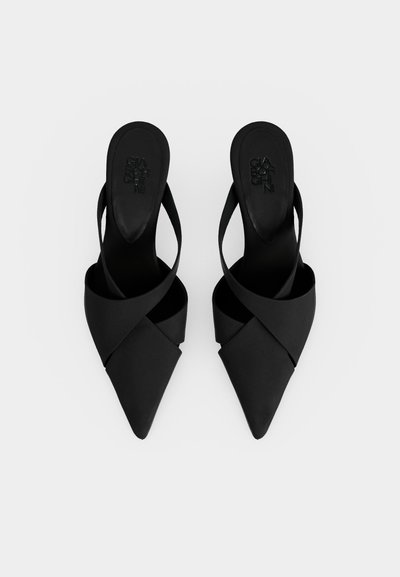 Par sorte spidse højhælede mule med krydsede stropper og åbne hæle på en ens hvid baggrund.