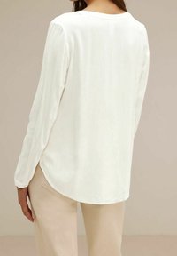 Blusa bianca a maniche lunghe con scollatura rotonda e orlo leggermente curvo. Tessuto liscio e leggero con una vestibilità morbida e un leggero lucido.