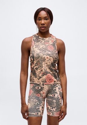 Femme athlétique debout, portant un haut sans manches et un short de cycliste assortis à motifs floraux et ondulés dans des tons beige, noir et rose.