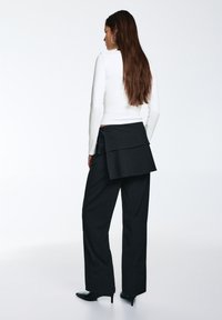 Pantalon noir à jambes larges avec un design superposé à l'arrière et un détail noué, associé à un haut blanc à manches longues. Tissu lisse, silhouette élégante.
