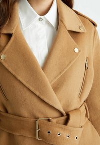 Dwurzędowy płaszcz z mieszanki wełny wielbłąda z srebrnymi okuciami, paskiem w talii i kieszeniami na zamek. W zestawie z białą koszulą z kołnierzykiem.